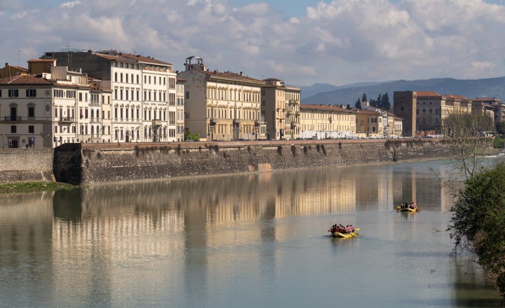 The Arno River reflecting Florence's white buildings along the edge. Two yellow rafts in the water with several people paddling on each of them.