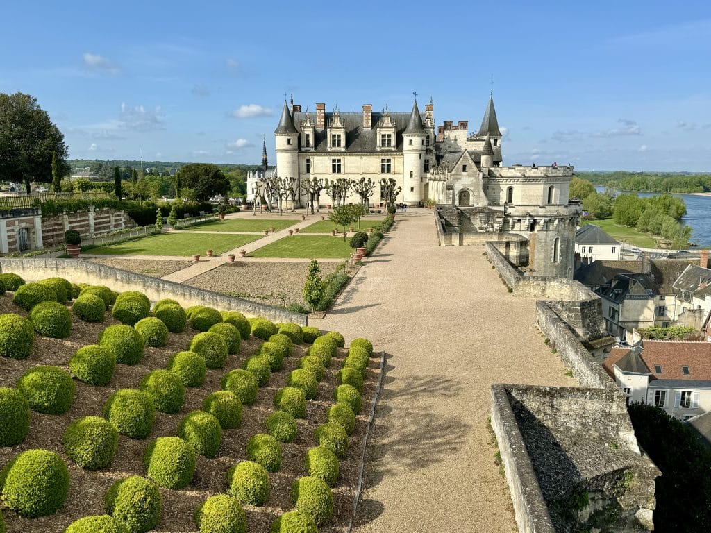 A gray castle in France perched on top of a hill, surrounded by nice landscaping, including rows of small green bushes.