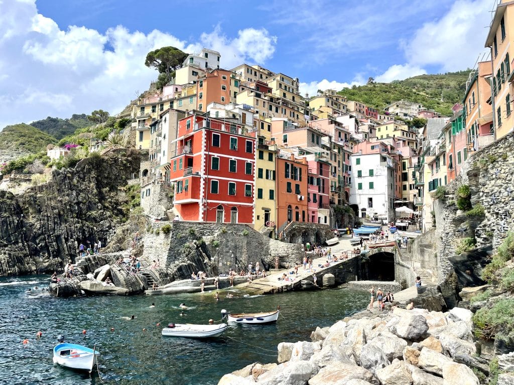 The town of Riomaggiore in Cinque Terre, all steep colorful buildings built into a rocky coastline above a blue-green sea strewn with rowboats.