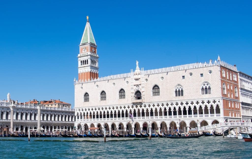 A palace on the edge of the water in Venice, white and tan colored with lots of white arches on the bottom floor. A pointy church tower rises behind it.