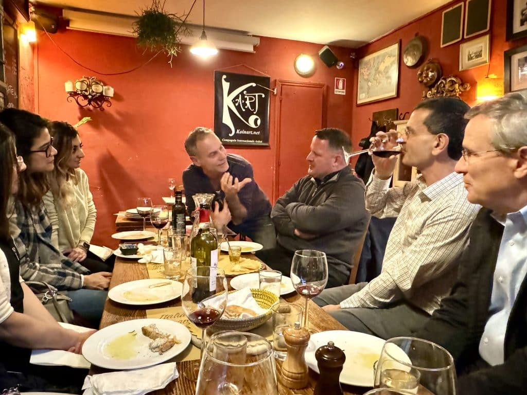 A group of people sitting around the table at a tiny restaurant in Italy as a guide leads a discussion.