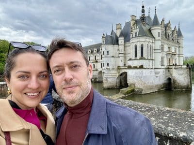 Kate and Charlie taking a smiling selfie in front of a big gray chateau perched on a river in France.