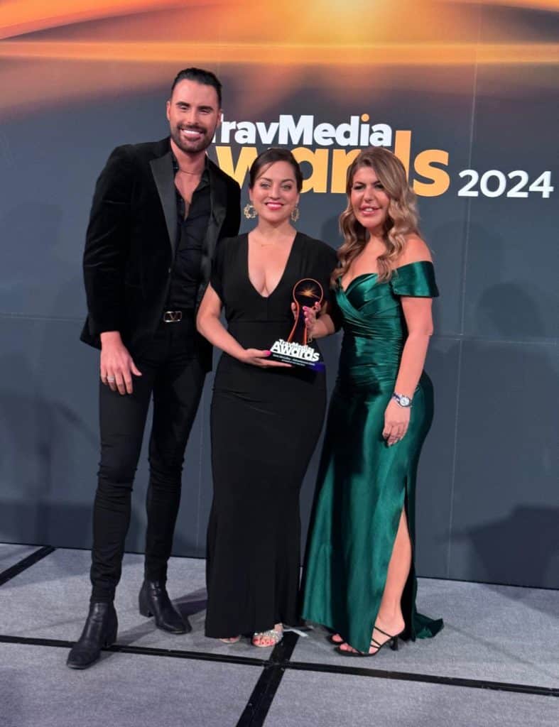 Kate in a long black evening gown holding up a trophy, standing between British reality celebrity Rylan (tall handsome man with a beard) and a woman in a long green gown.