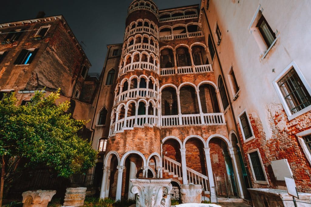 A Venetian palace with a unique covered spiral staircase on the outside, made of pink stone and wine trim.