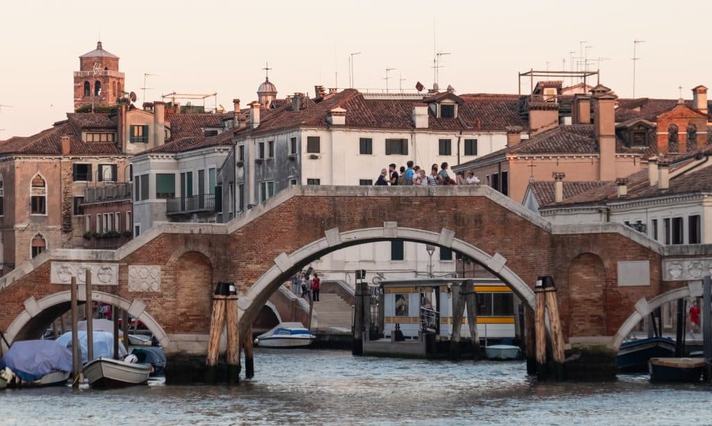 A curvy stone bridge over a canal in Venice, with a family standing on top, looking at the landscape around them.