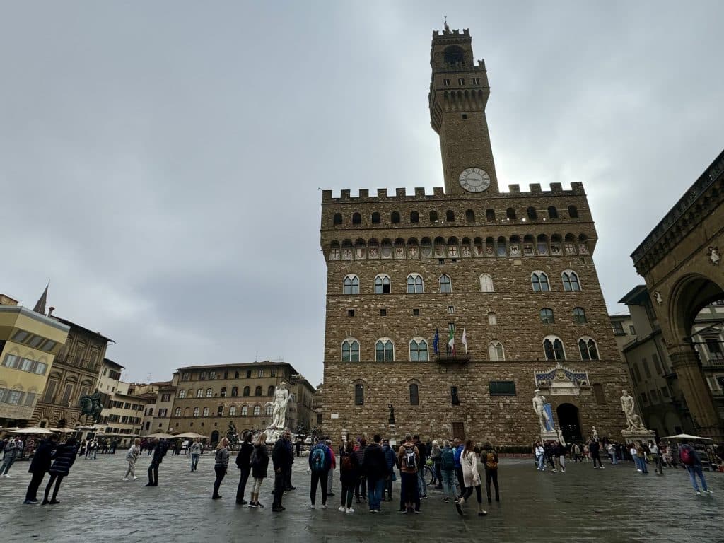 A tall palace with a stone tower on the edge of a big piazza in Florence, lots of tourists milling about.