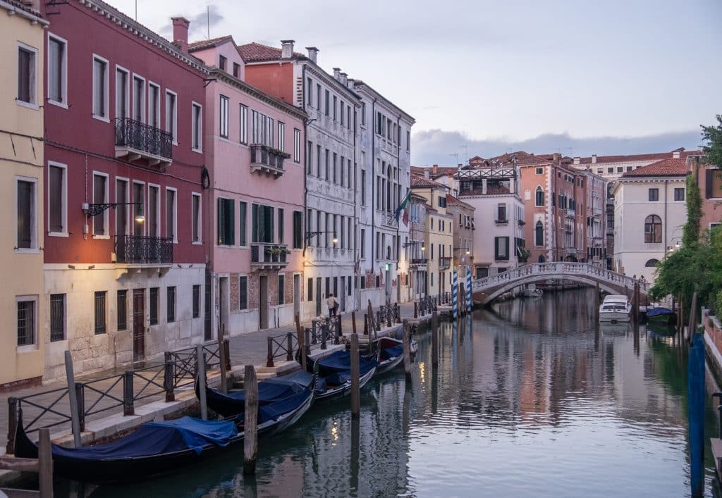 A quiet street giving way to a canal in Venice, the buildings all lit up in purple just after sunset, bridges crossing the calm waters.