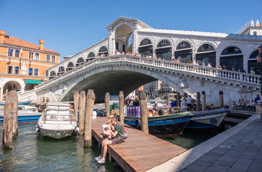 The big white Rialto bridge pointing above the Grand Canal, with a few people eating sandwiches on a dock in the foreground.