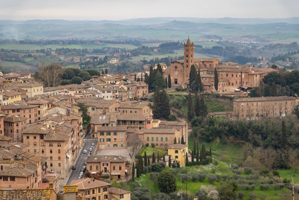 The Tuscan city of Siena, all made out of warm-toned brick with a church on a hill, surrounded by green hills.