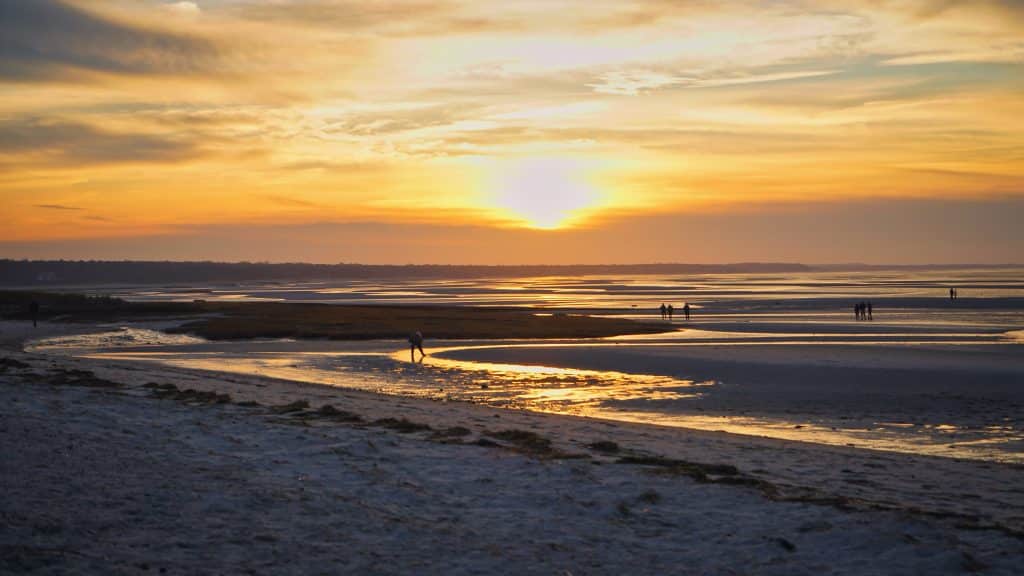 A Long Beach at low tide, underneath a golden sunset, lots of people walking along the beach.