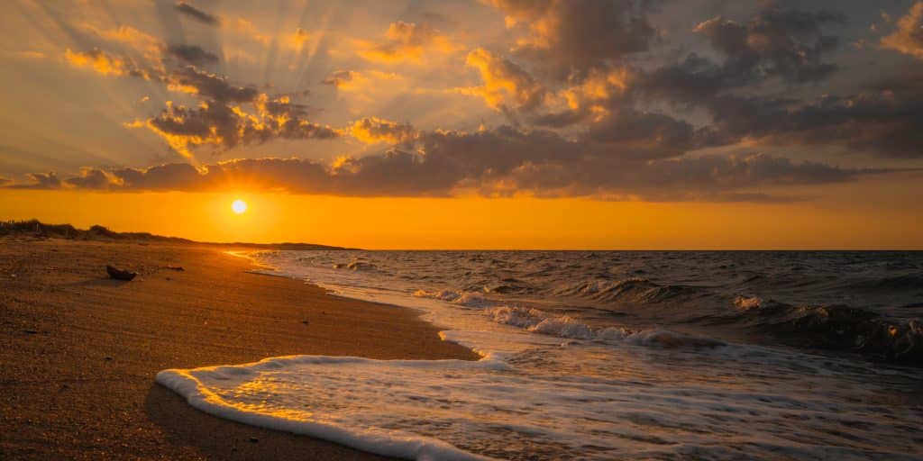 A bright orange and purple sunset as a wave cascades across a soft sand beach on Cape Cod.