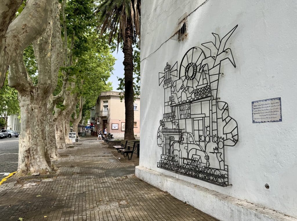 A sculpture of black metal twisted into the facade of a building with palm trees and a car in front, on a white building in Colonia.