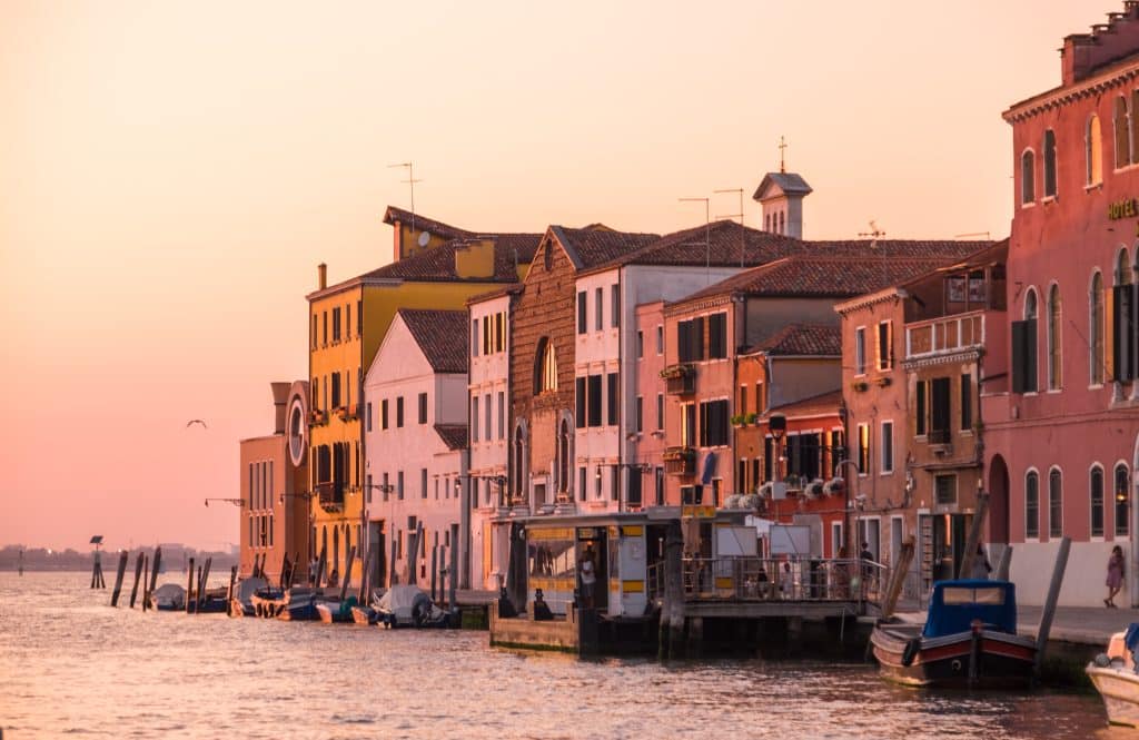 A long line of buildings on the edge of the canal lit up in bright pink sunset light.