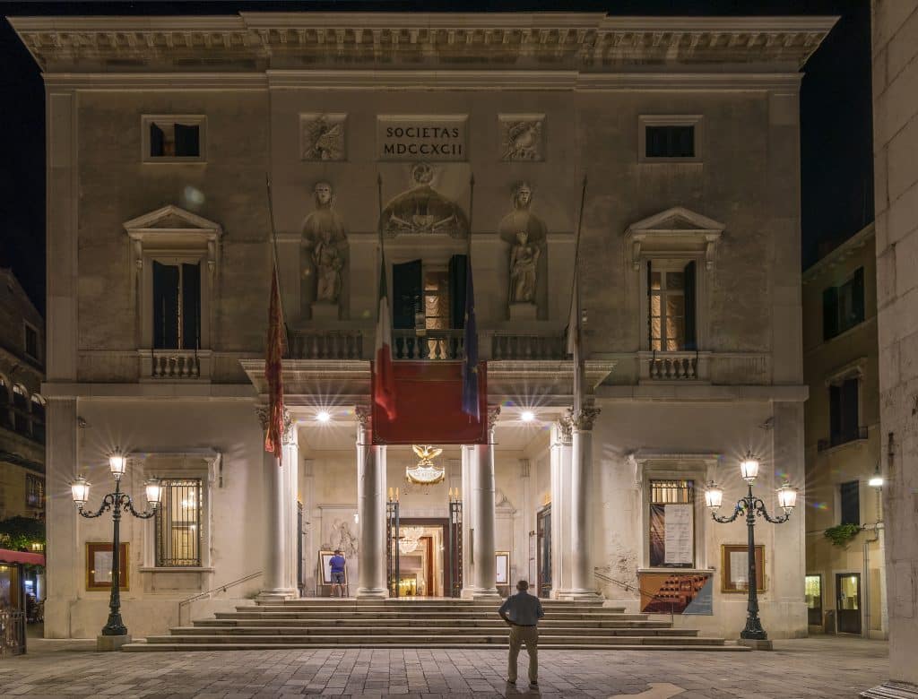 Venice's tall, white opera house, lit up from below at night, a man standing in front of it.