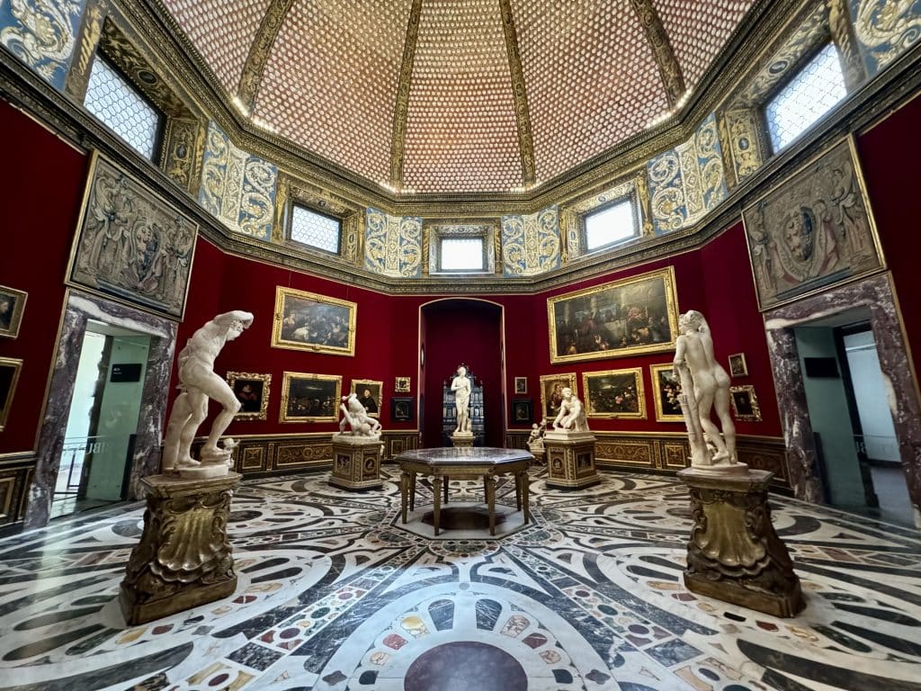 A bright red octangular room at the Uffizi featuring several marble statues, gold-framed paintings on the walls.