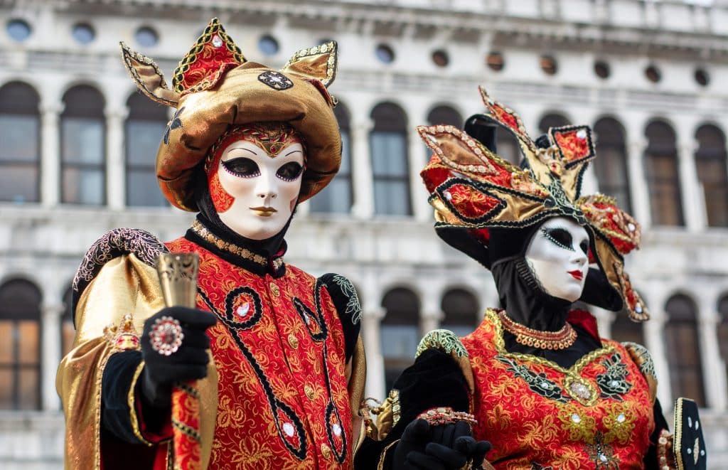 Performers in Venetian Masks and elaborate red and gold costumes, in front of a palace in Venice.