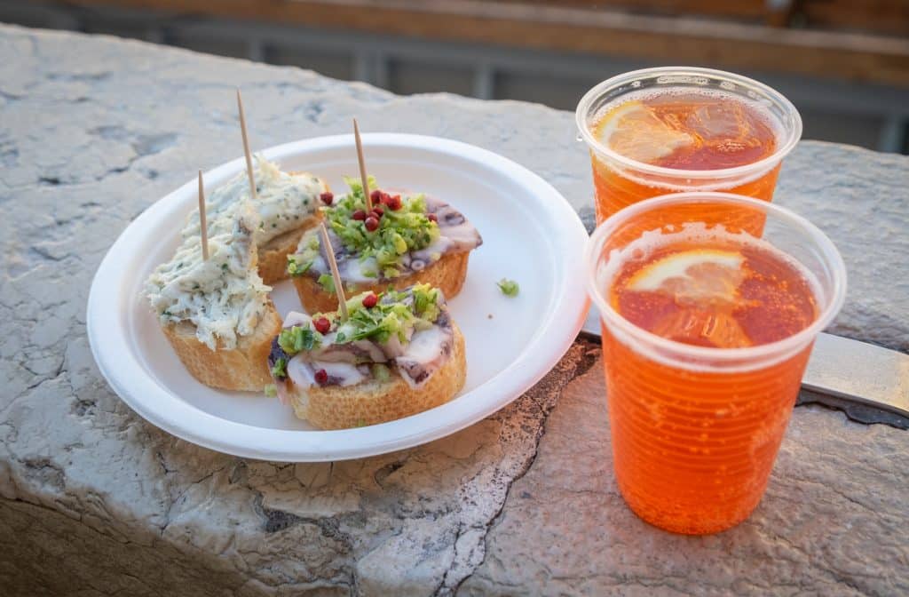 A paper plate with four little pieces of bread with toppings, next to two plastic cups full of aperol spritz.
