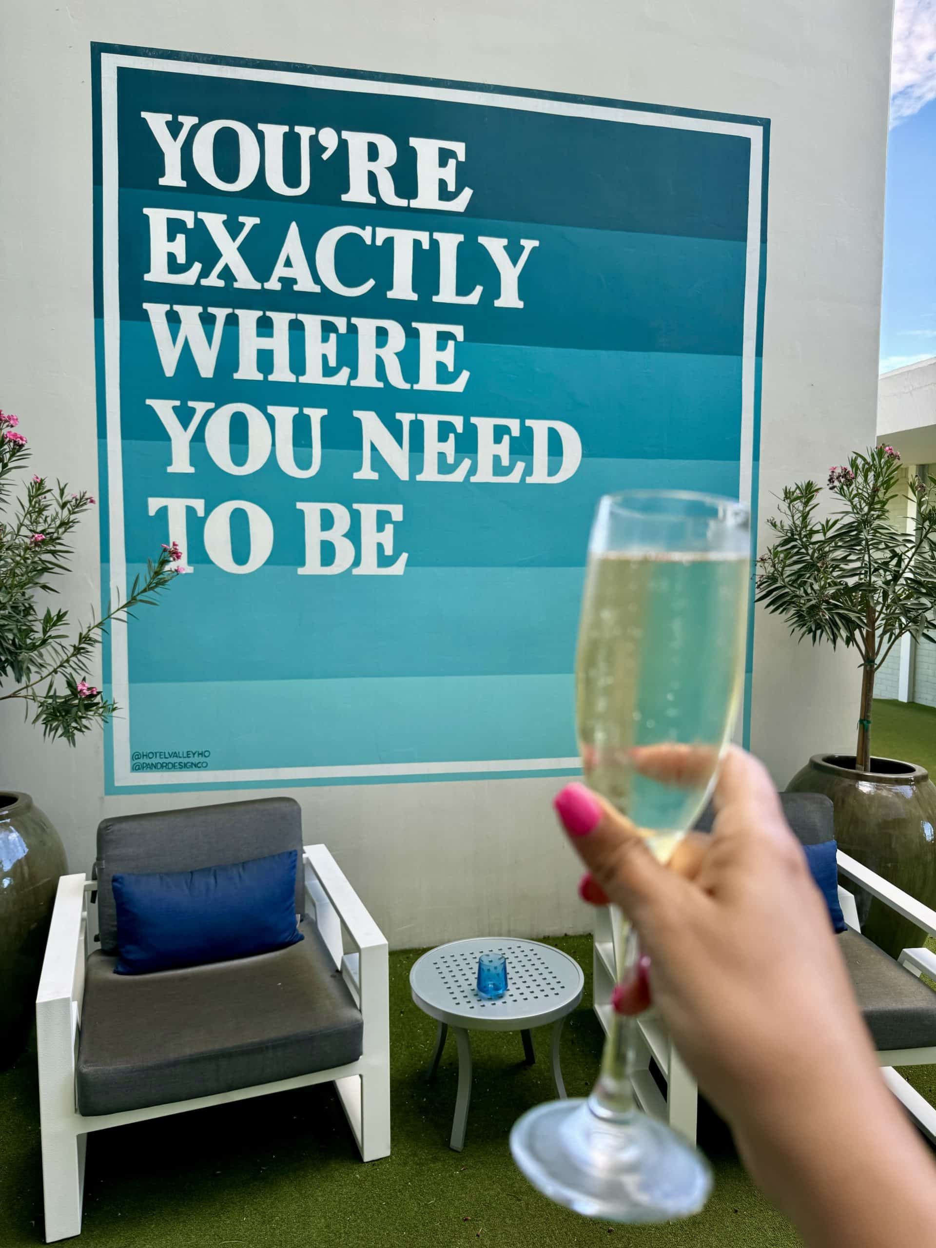 A glass of champagne held up in front of a mural reading "You're exactly where you need to be."