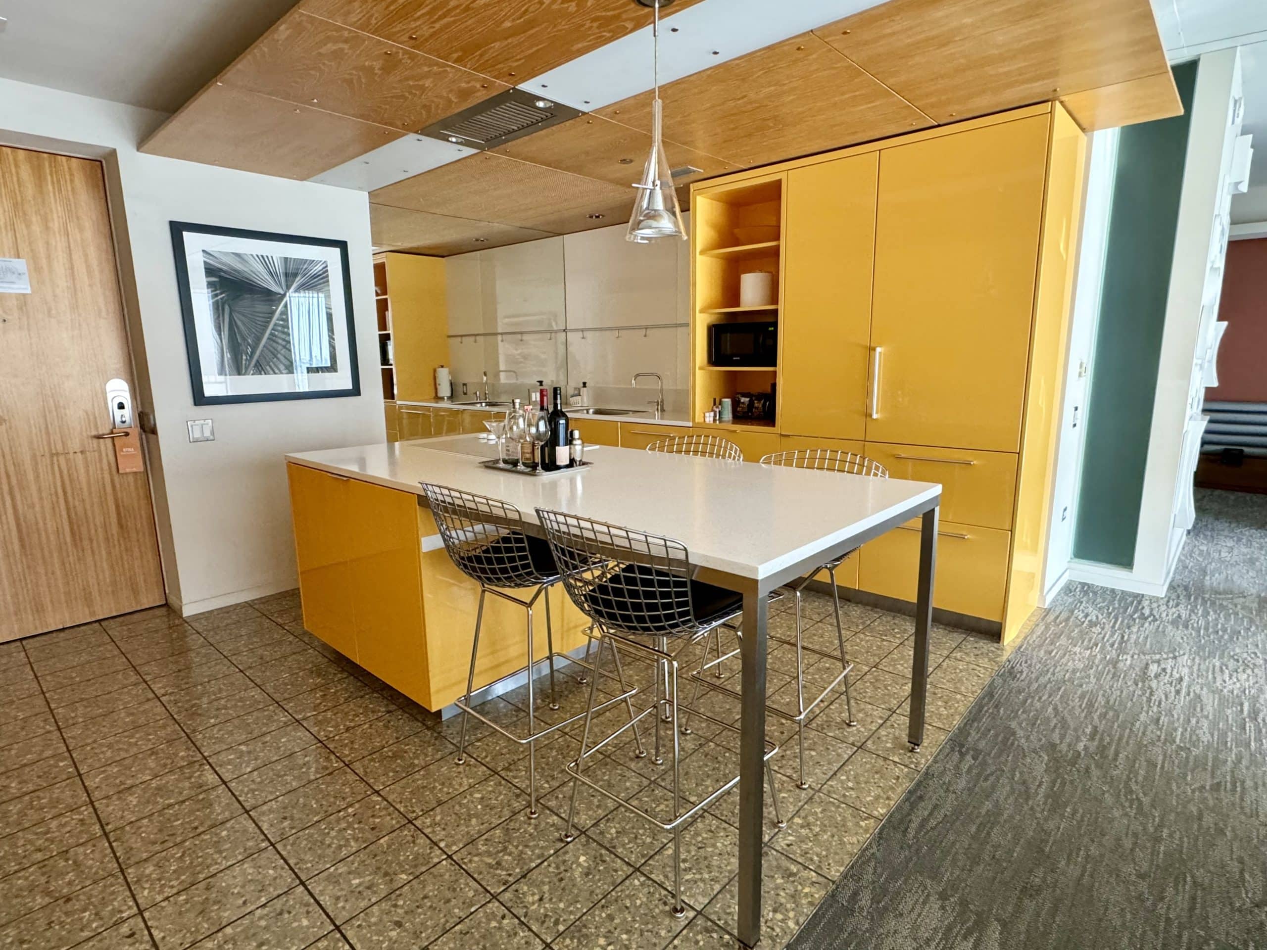 A hotel room kitchen with bright yellow cabinets, a sink, and two high-top chairs against the table.