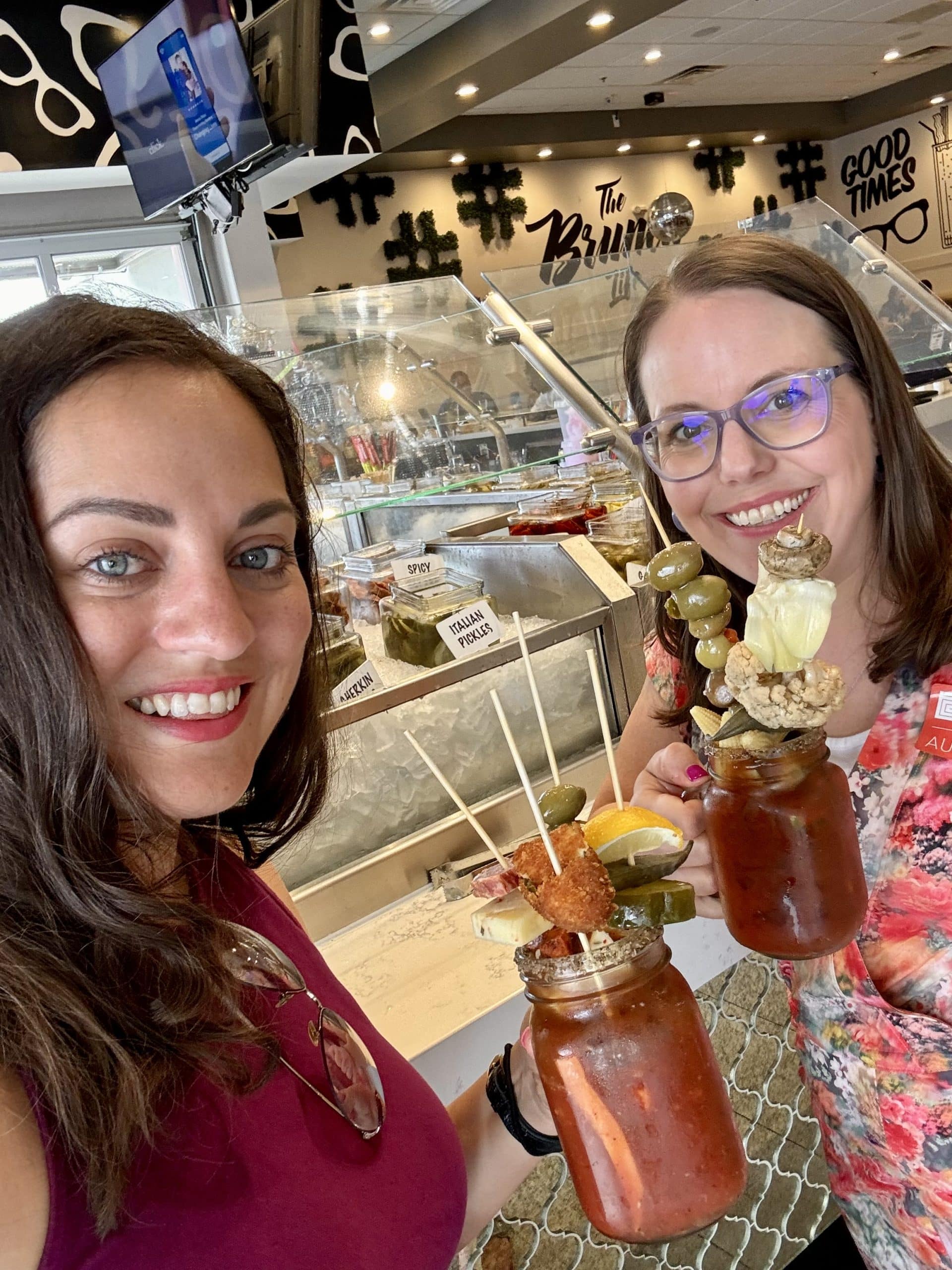 Kate and Beth holding up giant Bloody Marys covered with various pickled vegetables and accoutrements.