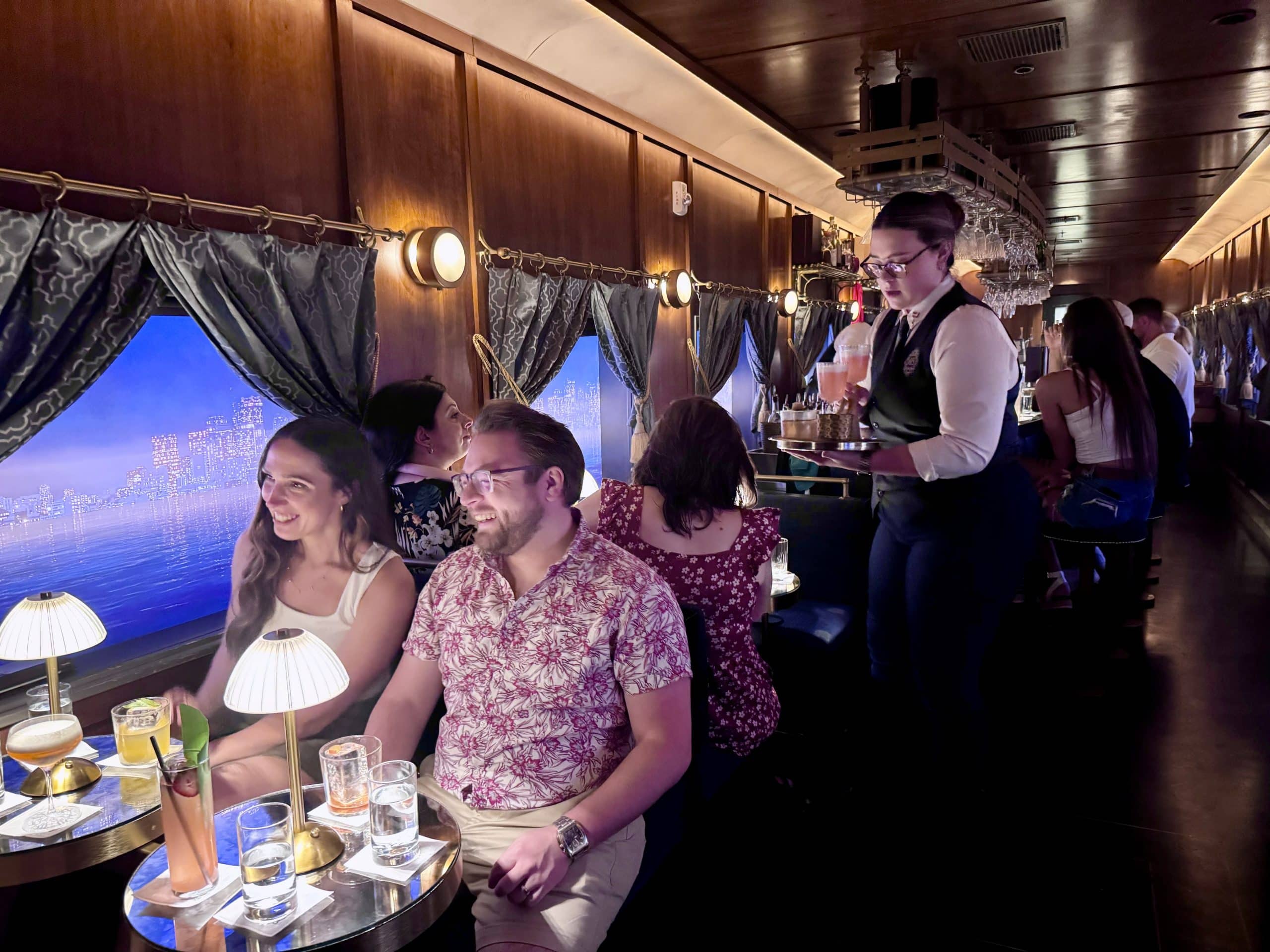 People sitting in a bar decorated like a 1920s train car, with a server in a waistcoat serving cocktails.
