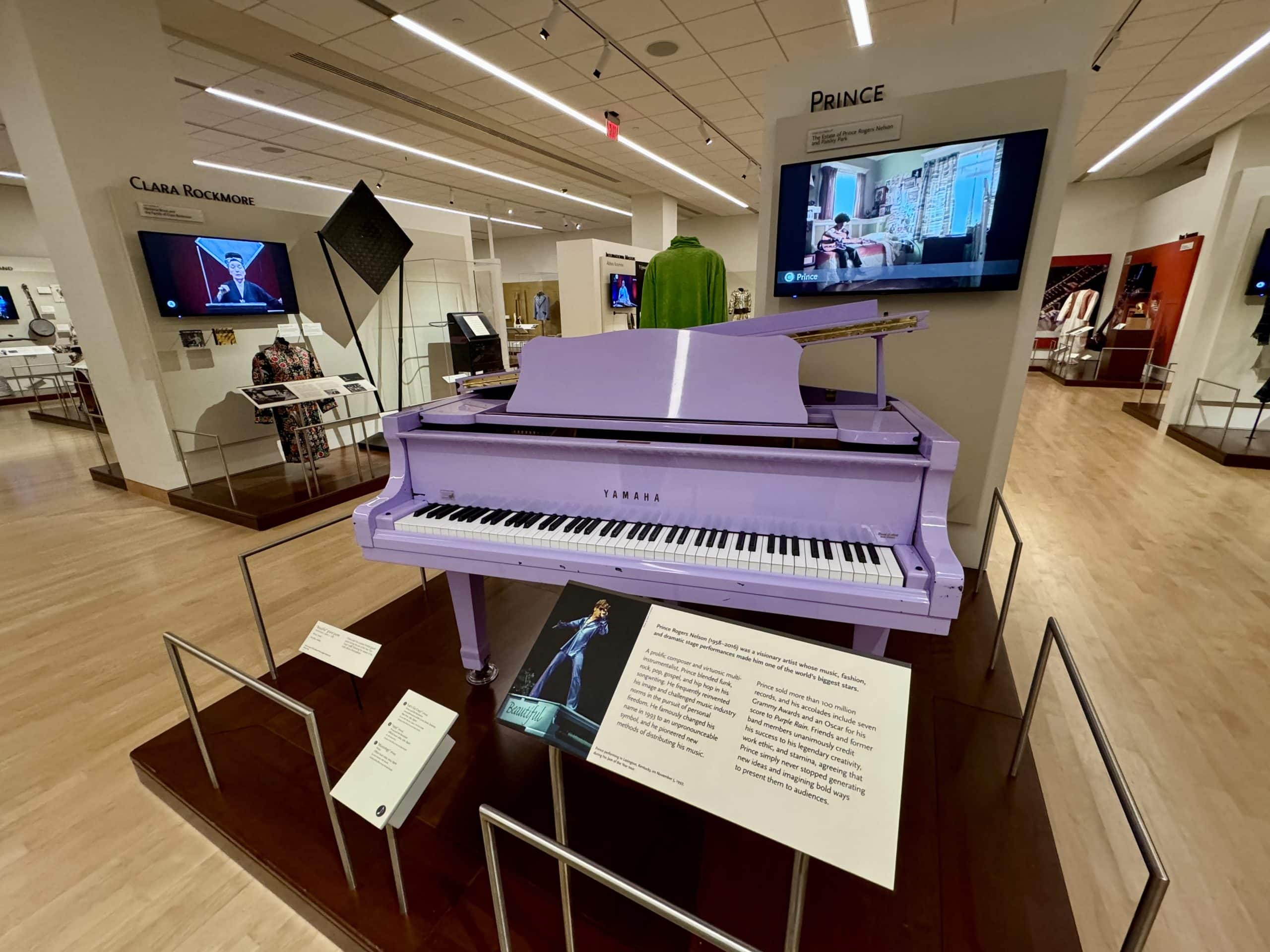A big purple piano on display in a museum, with a video of Prince on in the background.