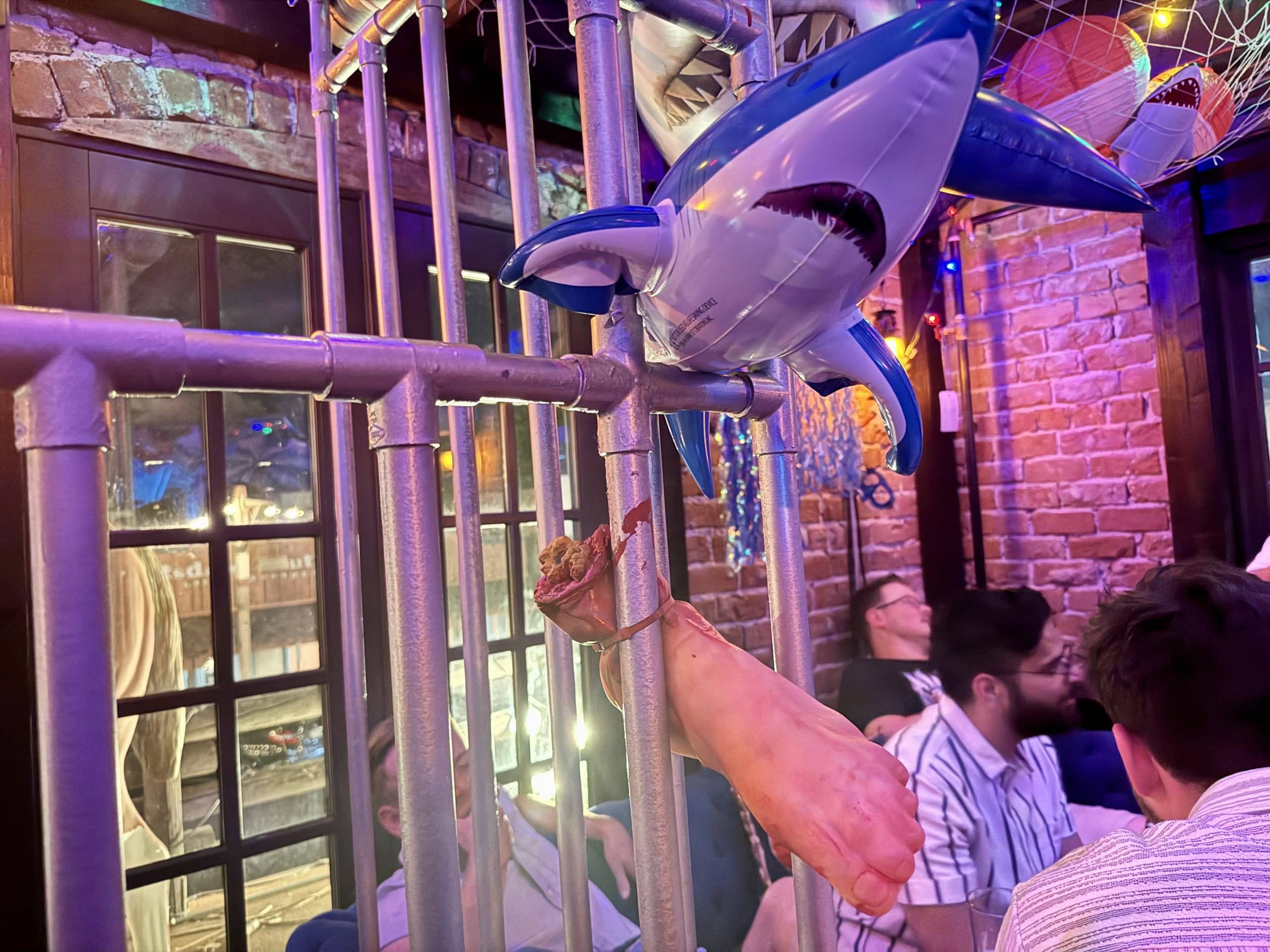 People hanging out in a bar, which has inflatable sharks and bloody limbs attached to a cage.