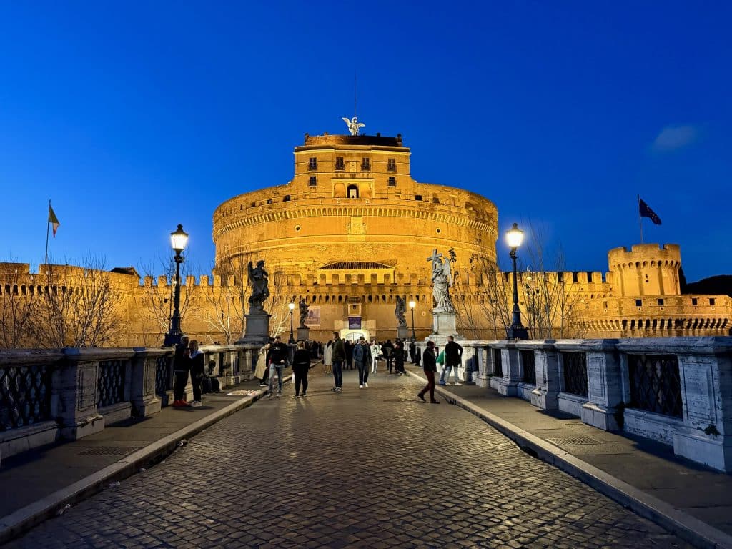 Rome's brick, round Castel Sant'Angelo lit up in orange against a deep blue sky after sunset.