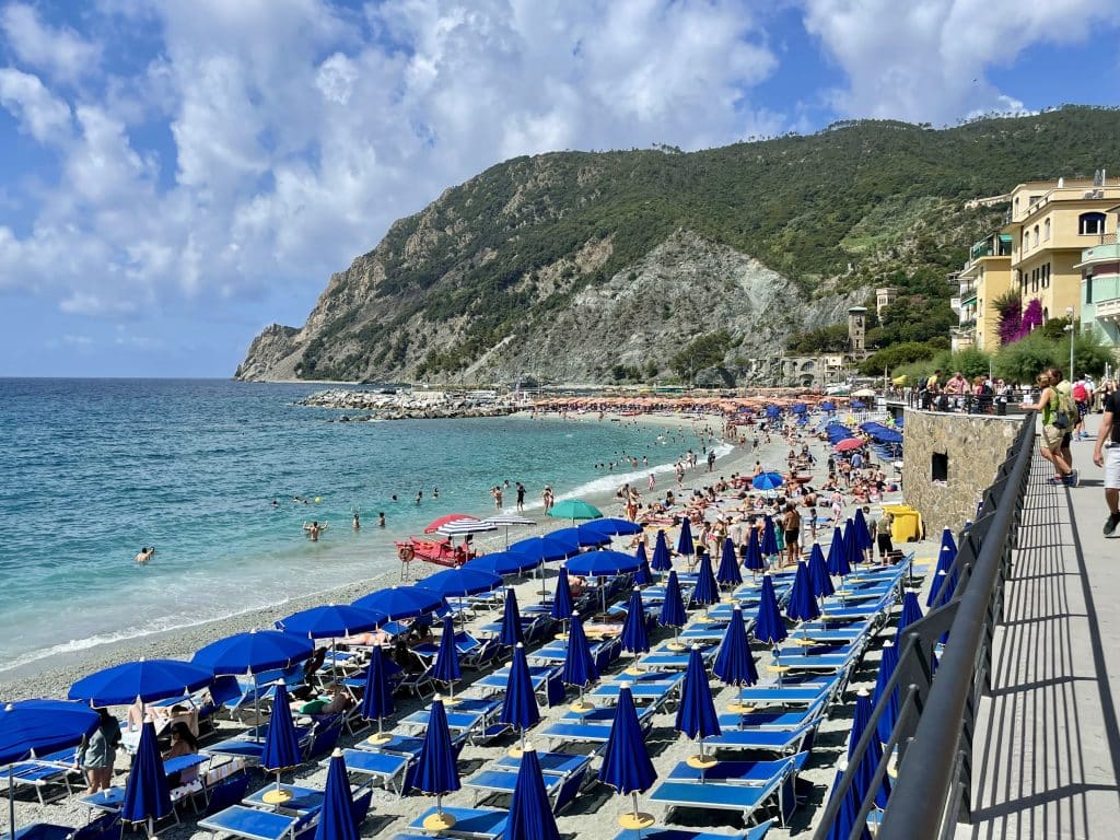 Rows of blue beach chairs and umbrellas on an Italian beach, with a big gray and green cliff in the background.