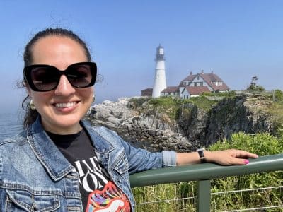 Kate taking a smiling selfie in sunglasses in front of a fog-surrounded lighthouse in Maine.