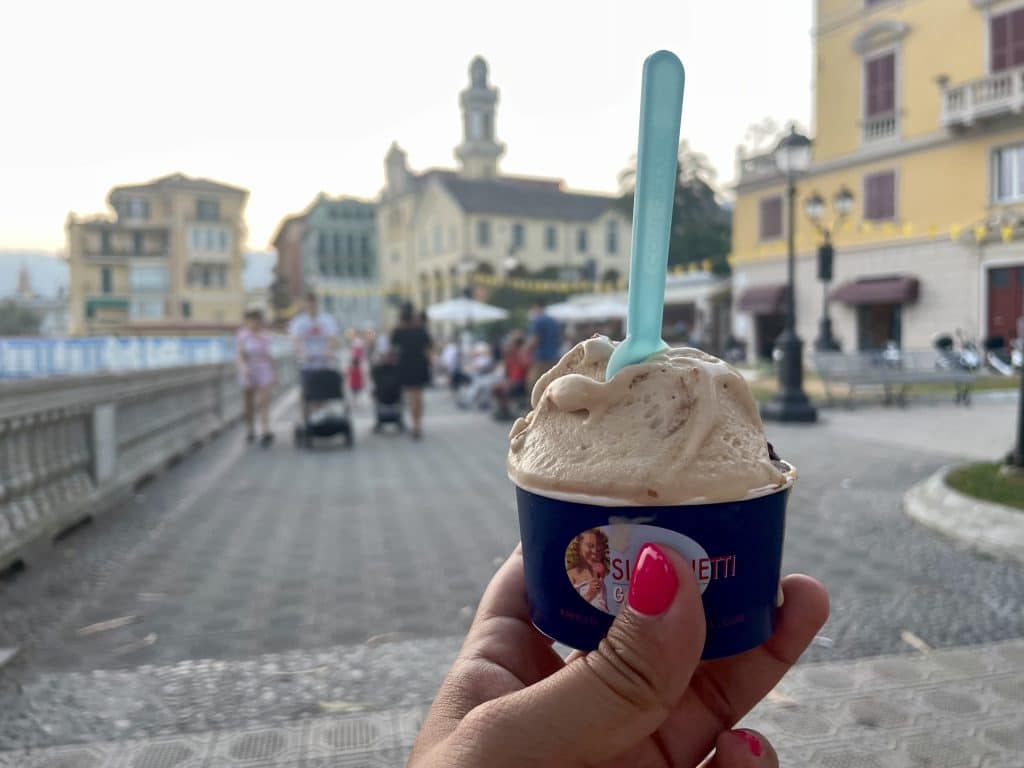 A hand holding up a cup of gelato with a piazza and church in the background.