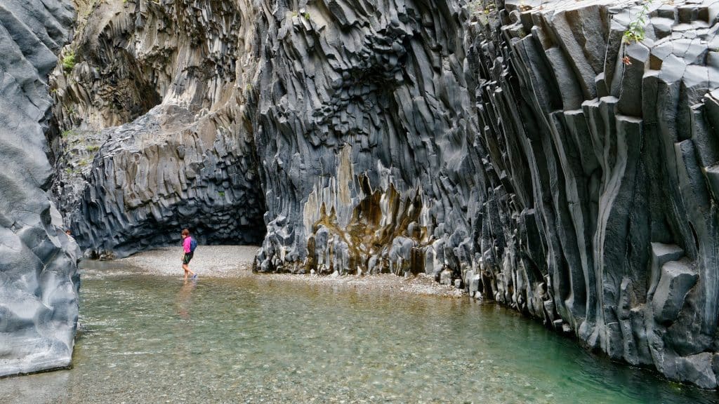 A woman backpacking through the bottom of a gorge, walking along a beach, surrounded by the gorge walls of basalt columns.
