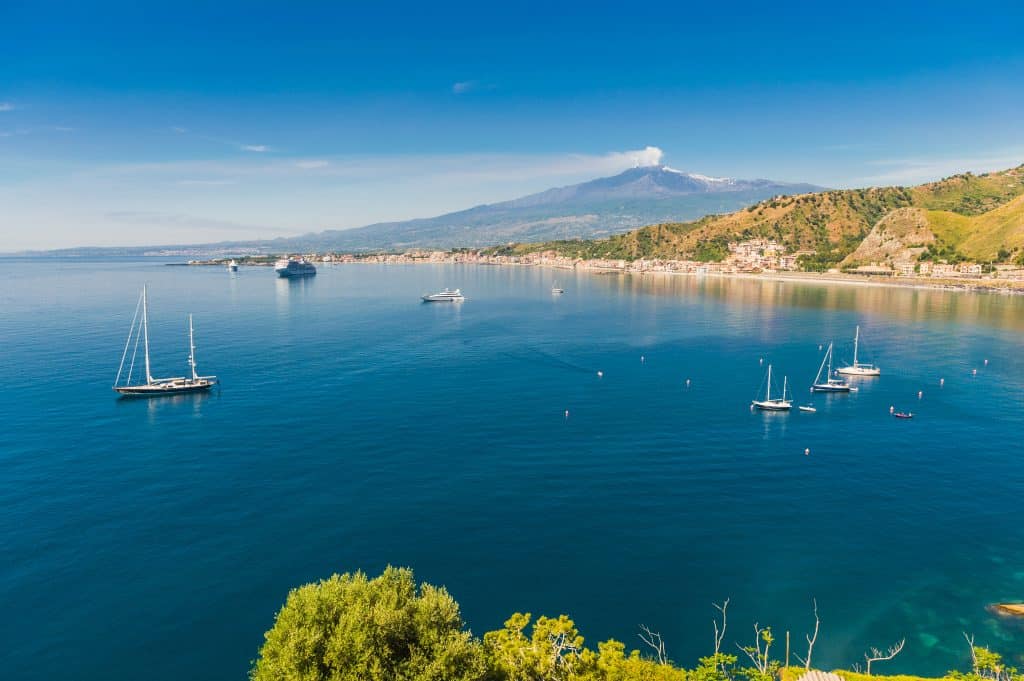 A bright blue sea and the coast of Sicily, with Mount Etna in the background.
