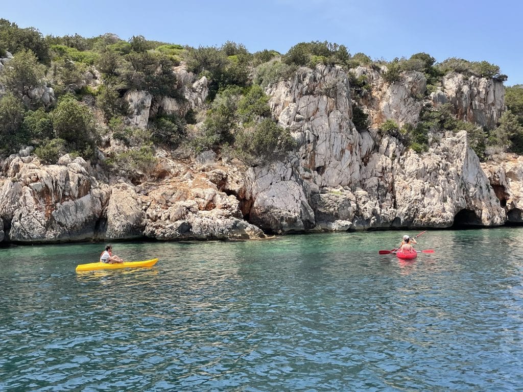 Two kayakers in yellow and red kayaks in a peaceful cove with cliffs and clear teal water.