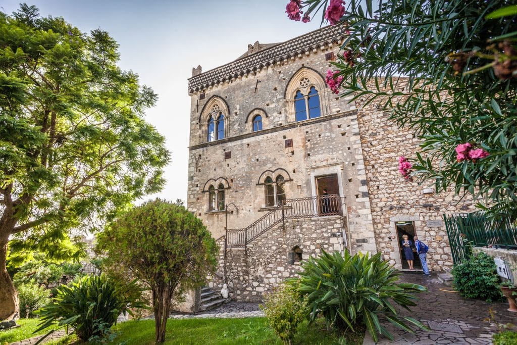 A stone palace in Sicily surrounded by lush greenery.