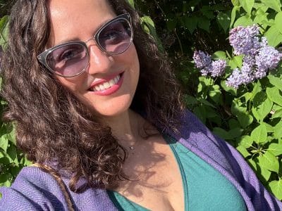 Kate smiling in sunglasses, standing in front of a lilac bush, wearing a green dress, purple shawl, and purple belt.