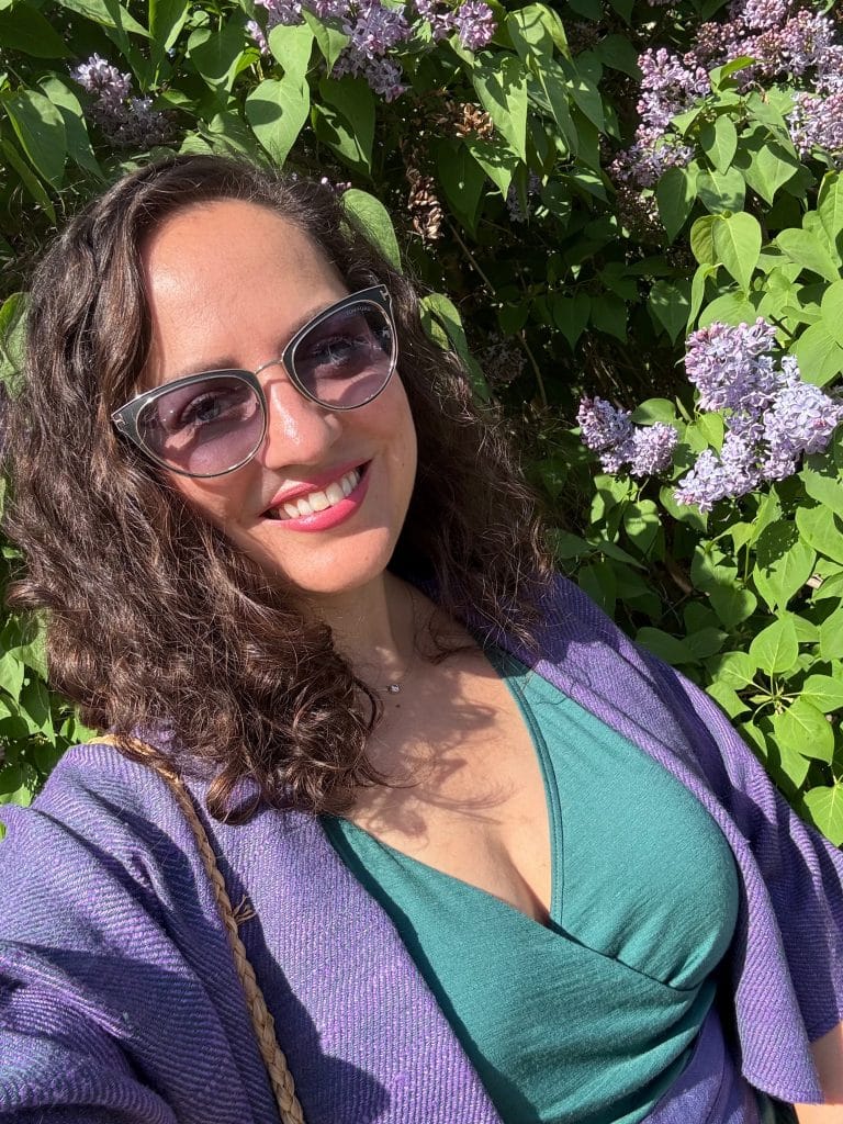 Kate smiling in sunglasses, standing in front of a lilac bush, wearing a green dress, purple shawl, and purple belt.