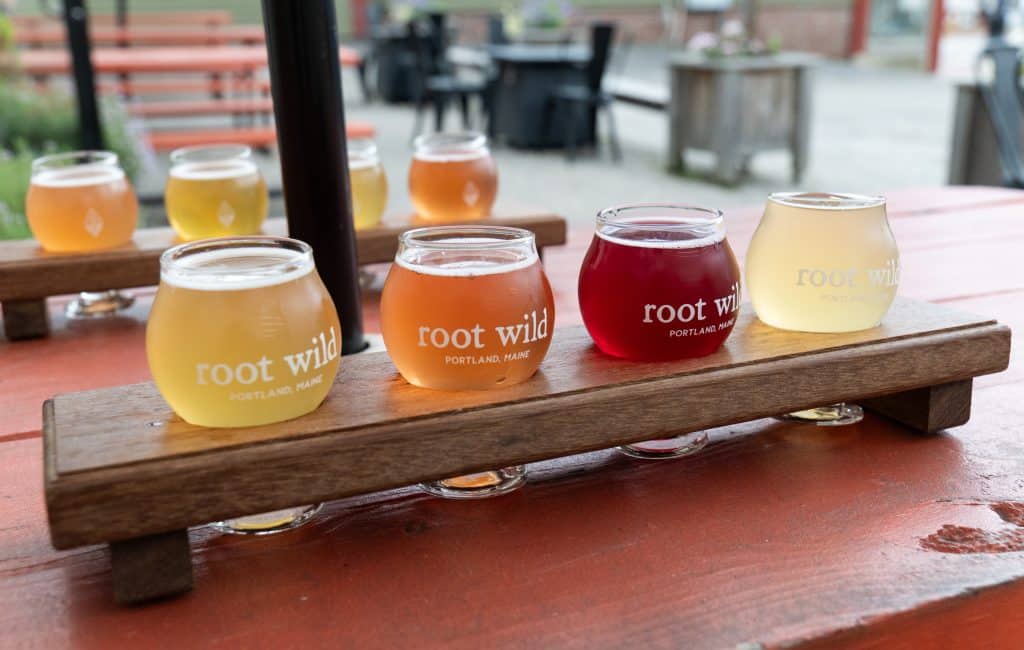 Four tasting glasses of kombucha labeled "Root Wild" on a wooden board.