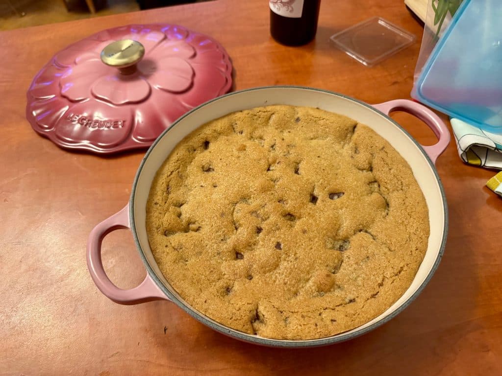 A giant cookie in a cast-iron skillet, with its bright pink flower-shaped lid to the side.