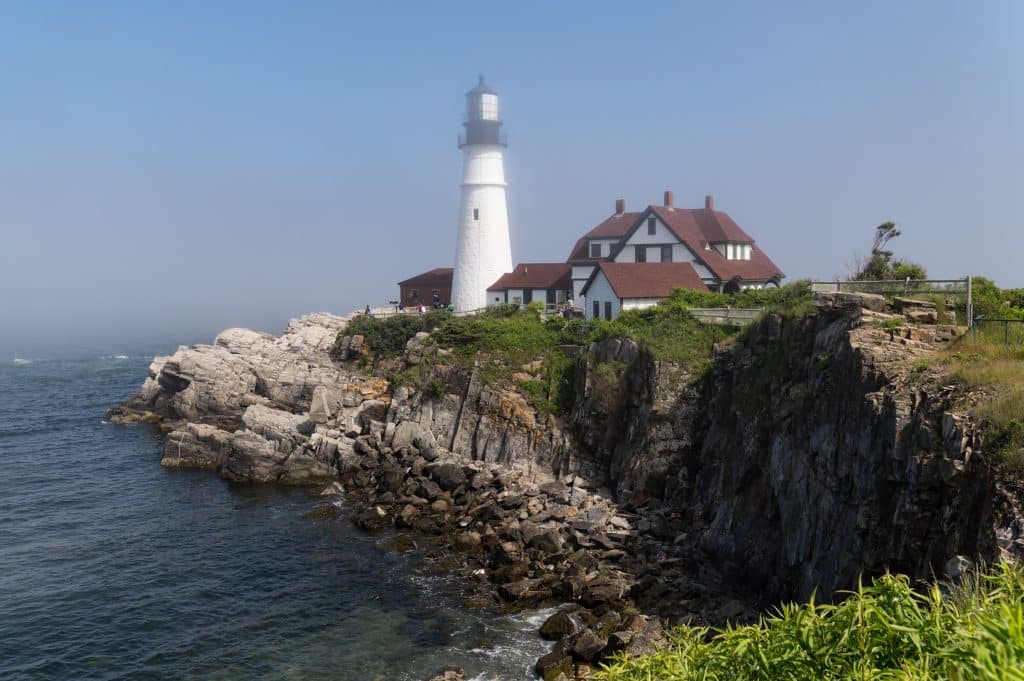 A huge white lighthouse perched on the rocky edge of the sea, topped with light fog.