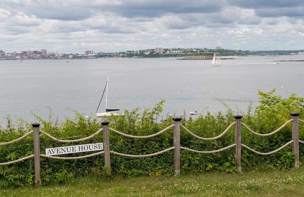 A view of Portland city across the water, with a small rope fence in front of greenery in the foreground. Boats are sailing in the water.