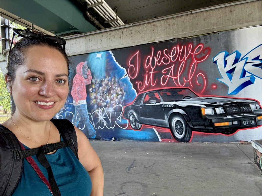 Kate taking a selfie in front of a Kendrick Lamar mural with a GNX car and "I deserve it all" in script.