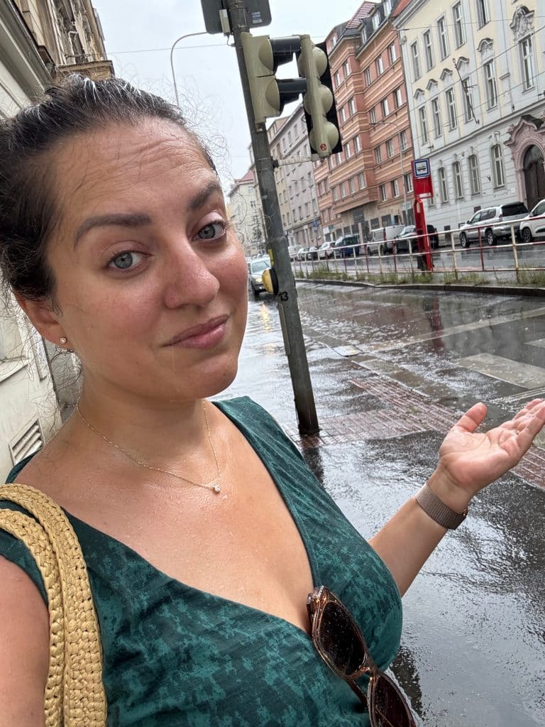 Kate standing on a rainy street, her green dress covered with raindrops, holding her hand up and shrugging.
