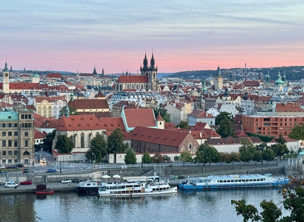The Prague skyline, with orange roofs and church steeples sticking up above. The sky is pink, just before sunset.
