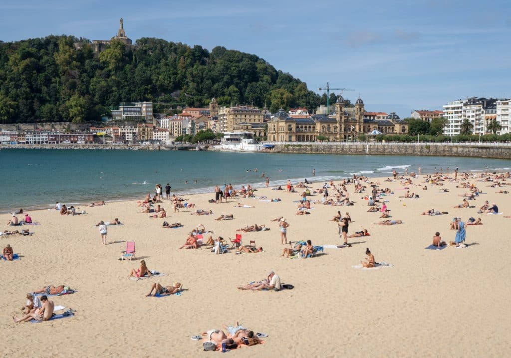 In the foreground, a beach with lots of sunbathers, and in the background, a big, tall, forested hill overlooking the water.