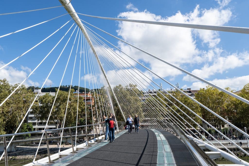 People walking along a bridge with tall white spires pointing upward along the edges.