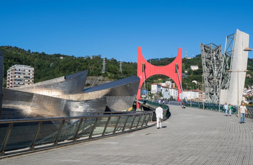 A bridge with a big red arc next to the shiny metal pieces of the Guggenheim Museum's roof.