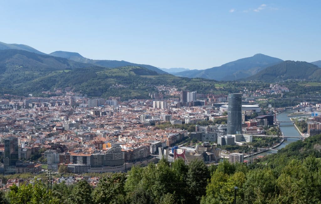A panoramic view of the city of Bilbao, with a skyscraper and the shiny, pointy Guggenheim museum in the foreground. There's a river running though the city and mountains in the background.