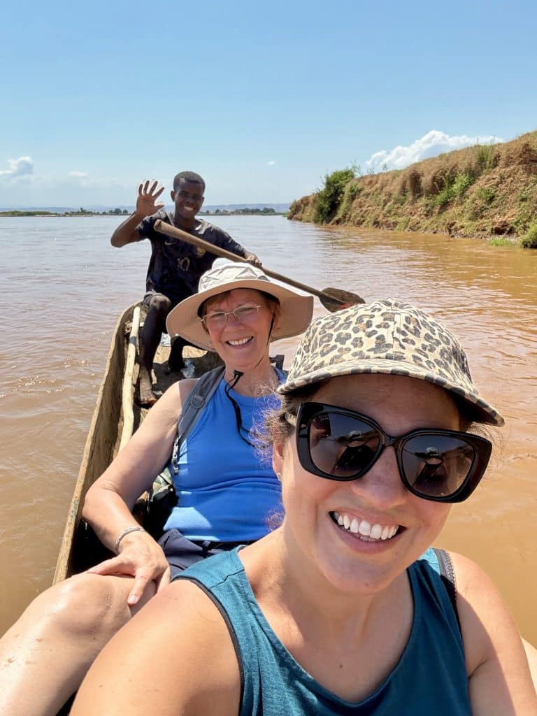 Kate and her friend Kim riding a canoe on a muddy river in Madagascar, the driver standing behind them and waving.