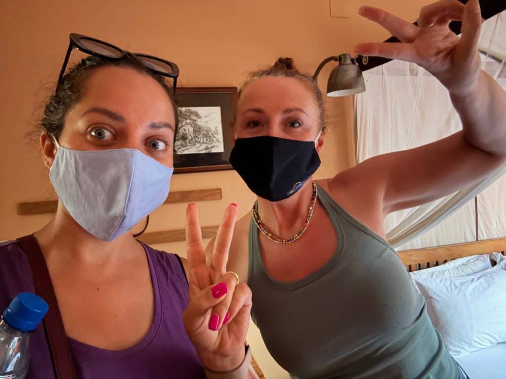 Kate and Helen wear cloth face masks and give peace signs within a hotel room.